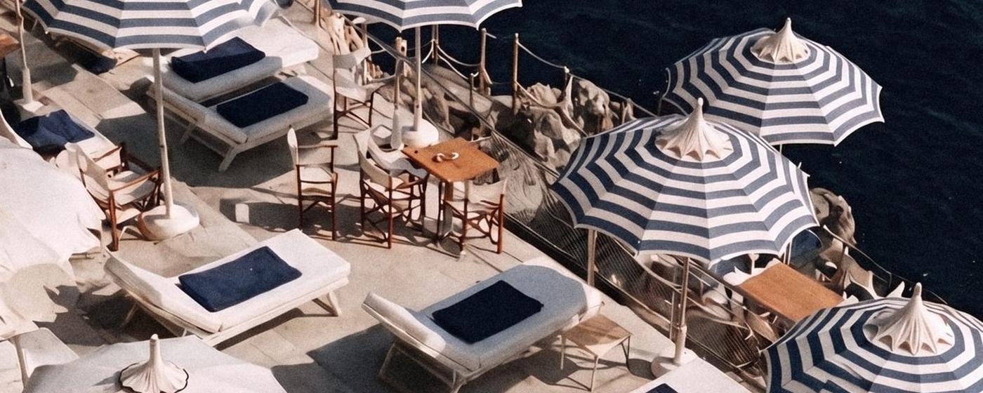 Beach club from above: striped umbrellas, white loungers, a wooden launch below